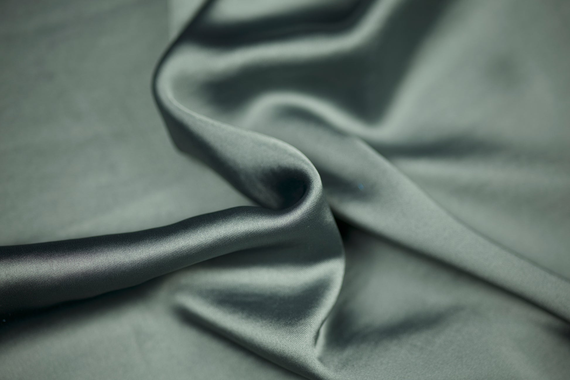 Nahaufnahme eines luxuriösen Satinstoffs in elegantem Salbeigrün mit sanftem Schimmer und natürlichem Faltenwurf – ideal für dezente Mode und Wohntextilien.
